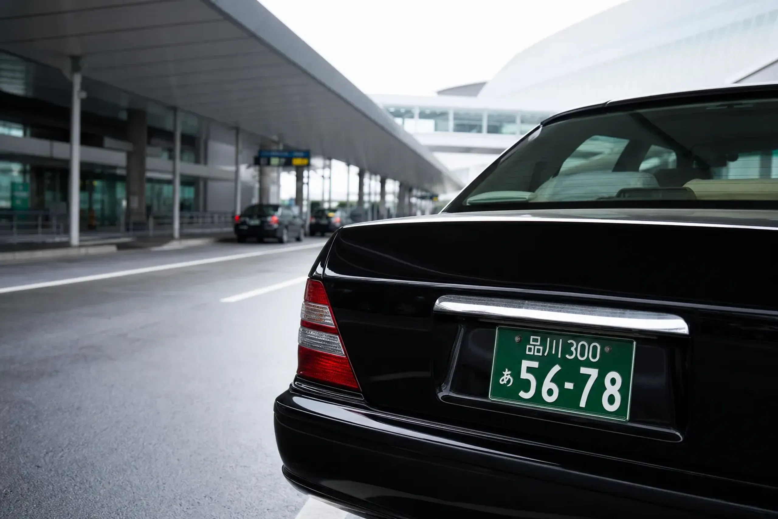 A green commercial passenger license plate on the rear of a black hire-car parked at a Tokyo airport curbside