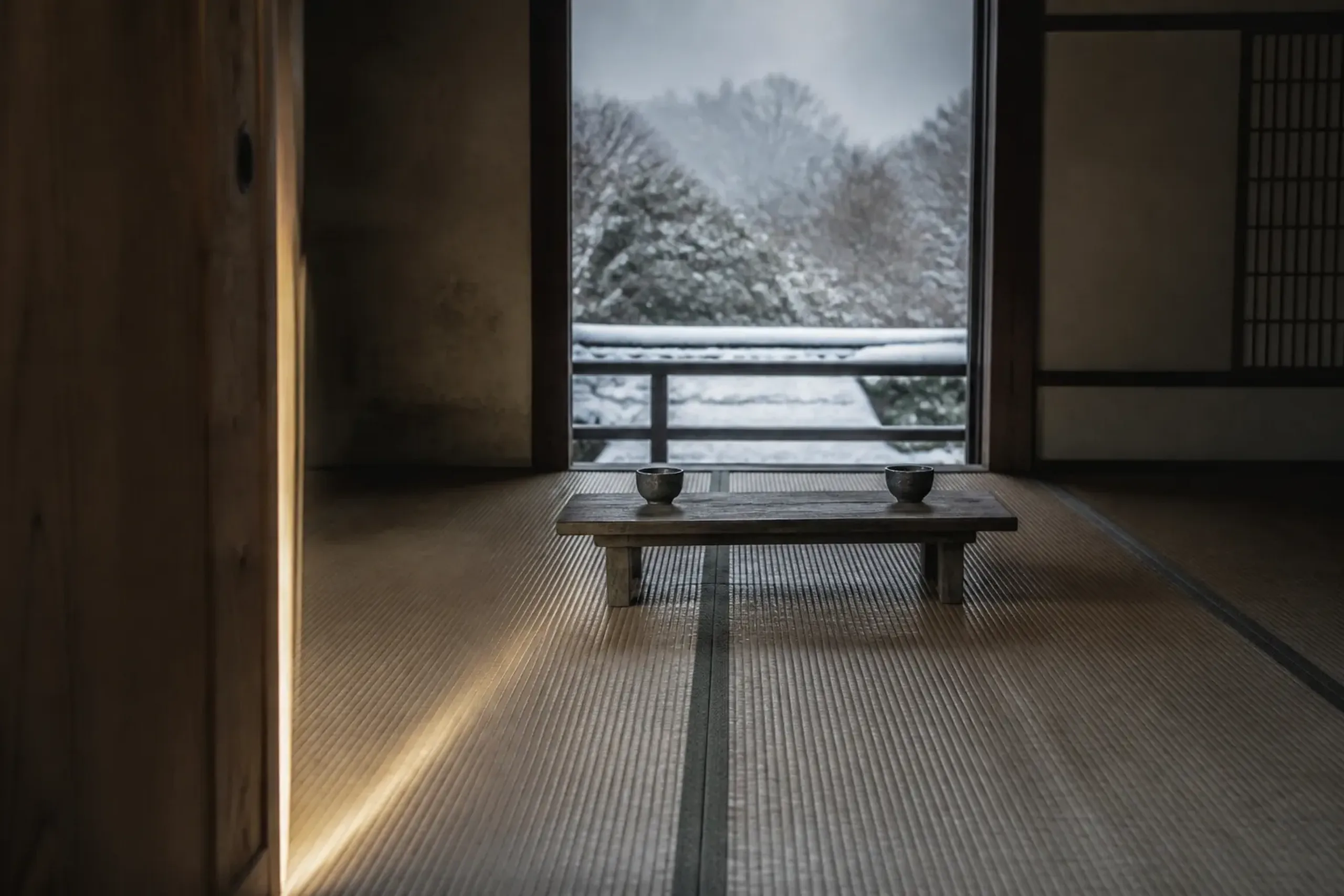 A quiet Kyoto artisan's atelier interior with an unadorned wooden sliding door left ajar, no signage or waiting-list board, a single low tea table with two cups set for an introductory meeting