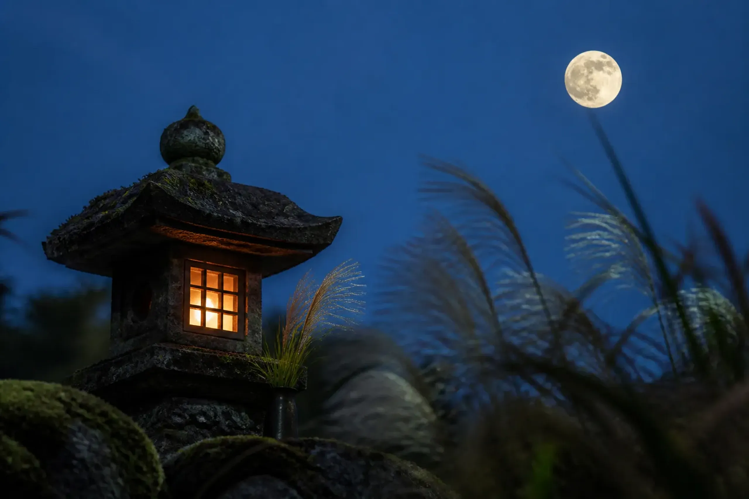 Stone lantern lit in a quiet Japanese garden with autumn grasses and a full moon rising