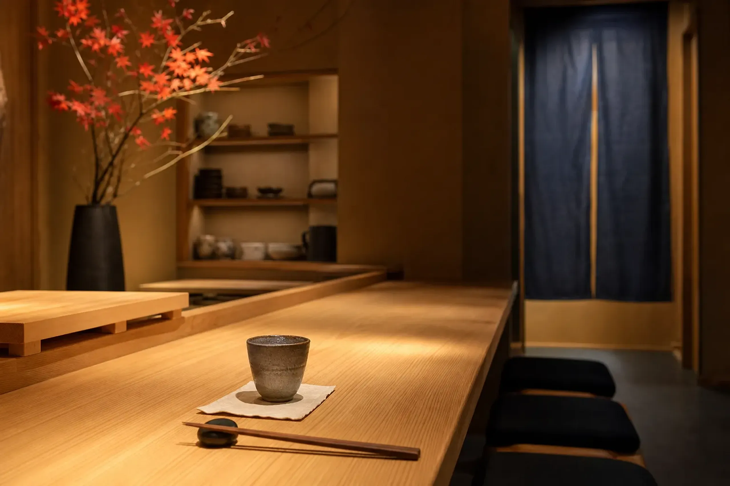 the dimly lit hinoki wood counter of an eight-seat sushi-ya in Kyoto, with a single ceramic sake cup set at a reserved place setting and a folded indigo noren curtain in the background