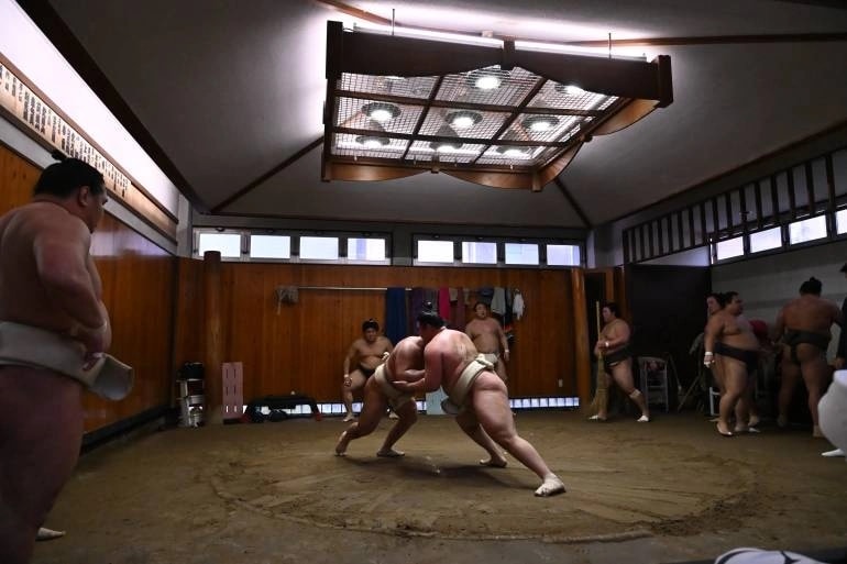 Two sumo wrestlers clash in a circular ring while others watch in a wooden dojo interior with a large ceiling light.