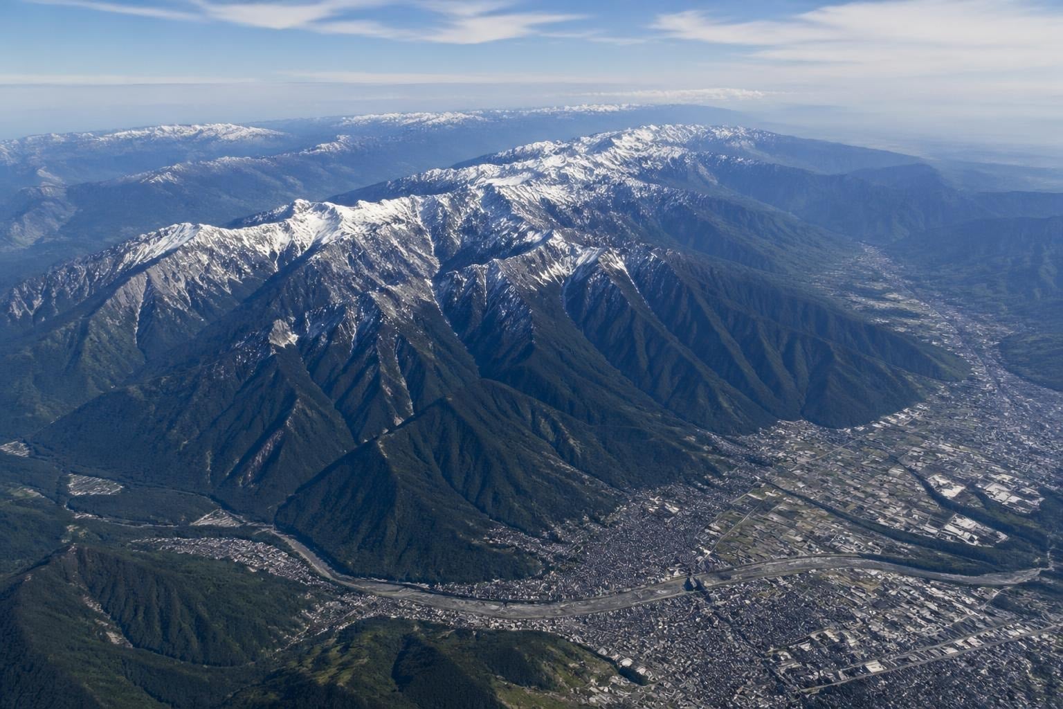 Aerial view of a snow-capped mountain range with a valley below, showing a winding river and a densely built urban area along the valley floor.