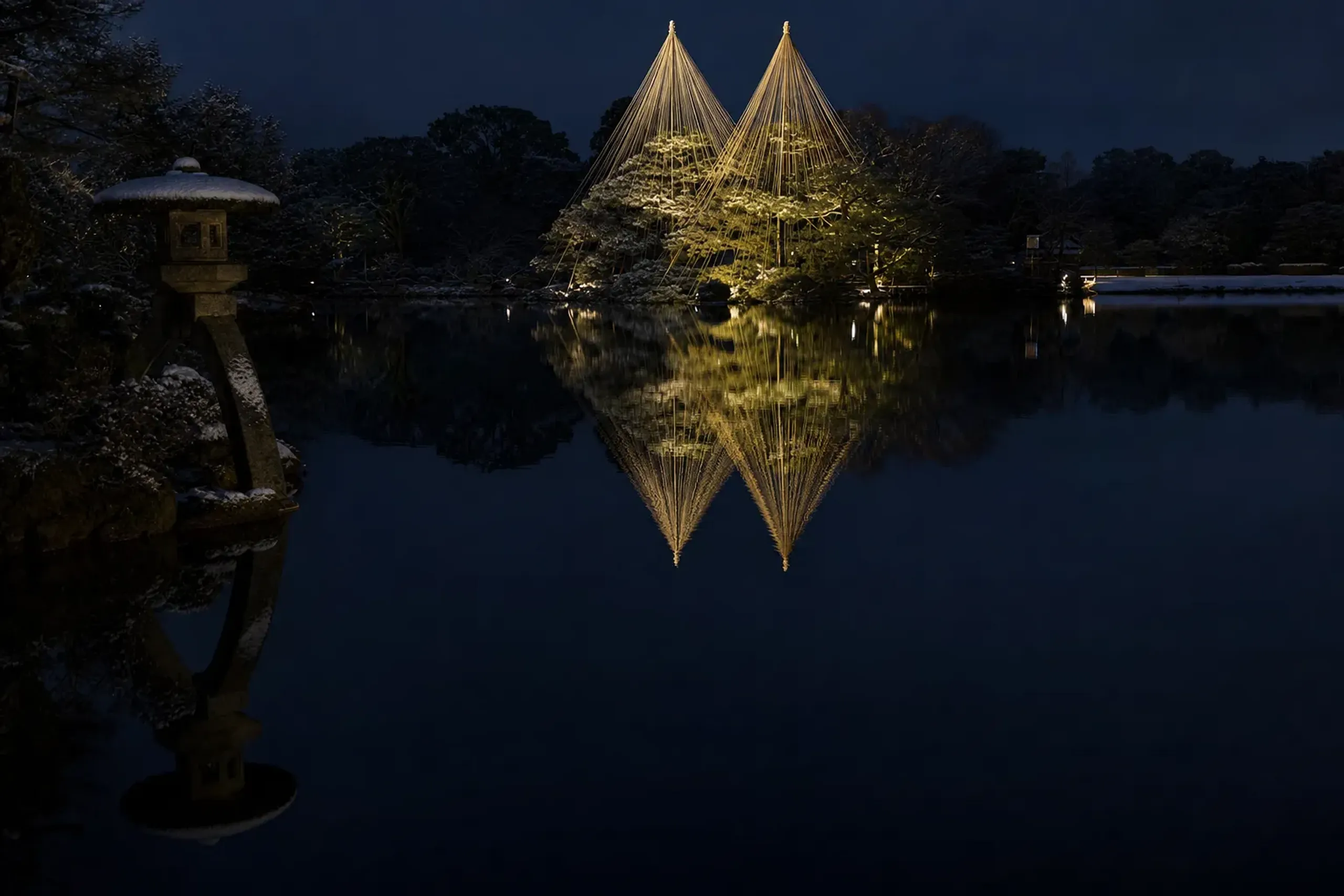 The illuminated golden yukitsuri rope cones reflected across the dark surface of Kasumigaike pond during Kenrokuen's February evening light-up