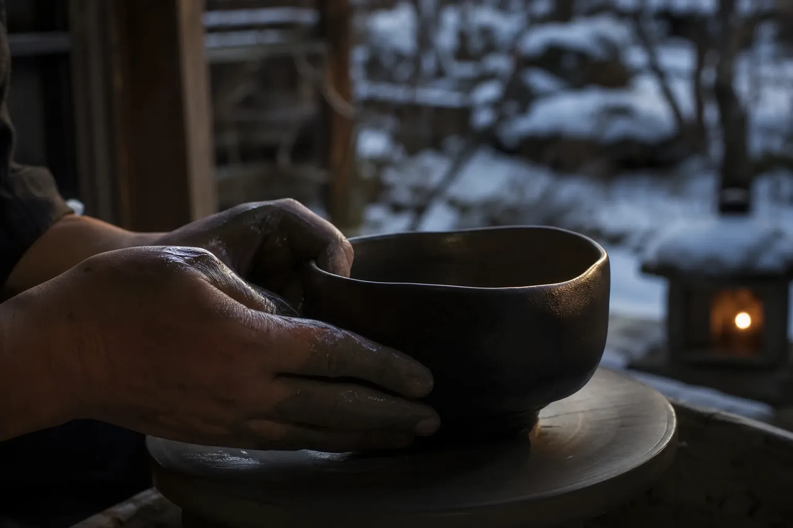 a hand-shaped Raku tea bowl with subdued dark glaze and deliberate uneven surface resting on a potter's wheel, wet clay and a maker's fingers mid-shaping
