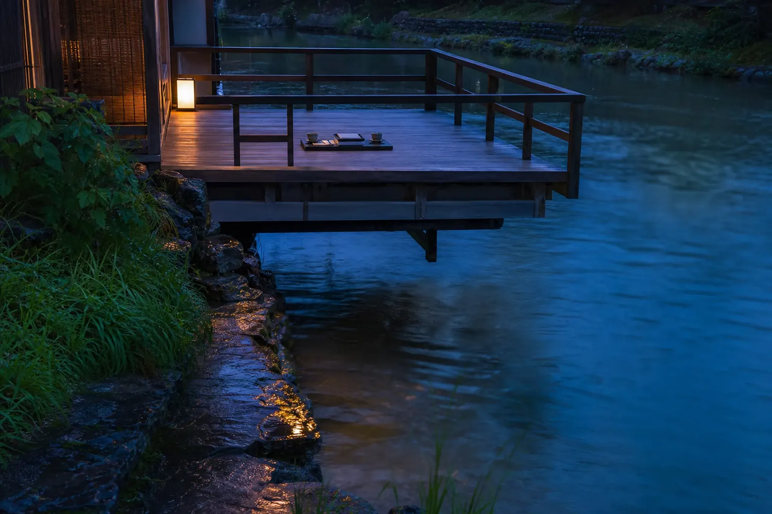 a private one-suite-per-day noryo-yuka wooden deck extending over the Kamogawa river at dusk, with an empty reserved dining setting and lantern light reflecting on the water