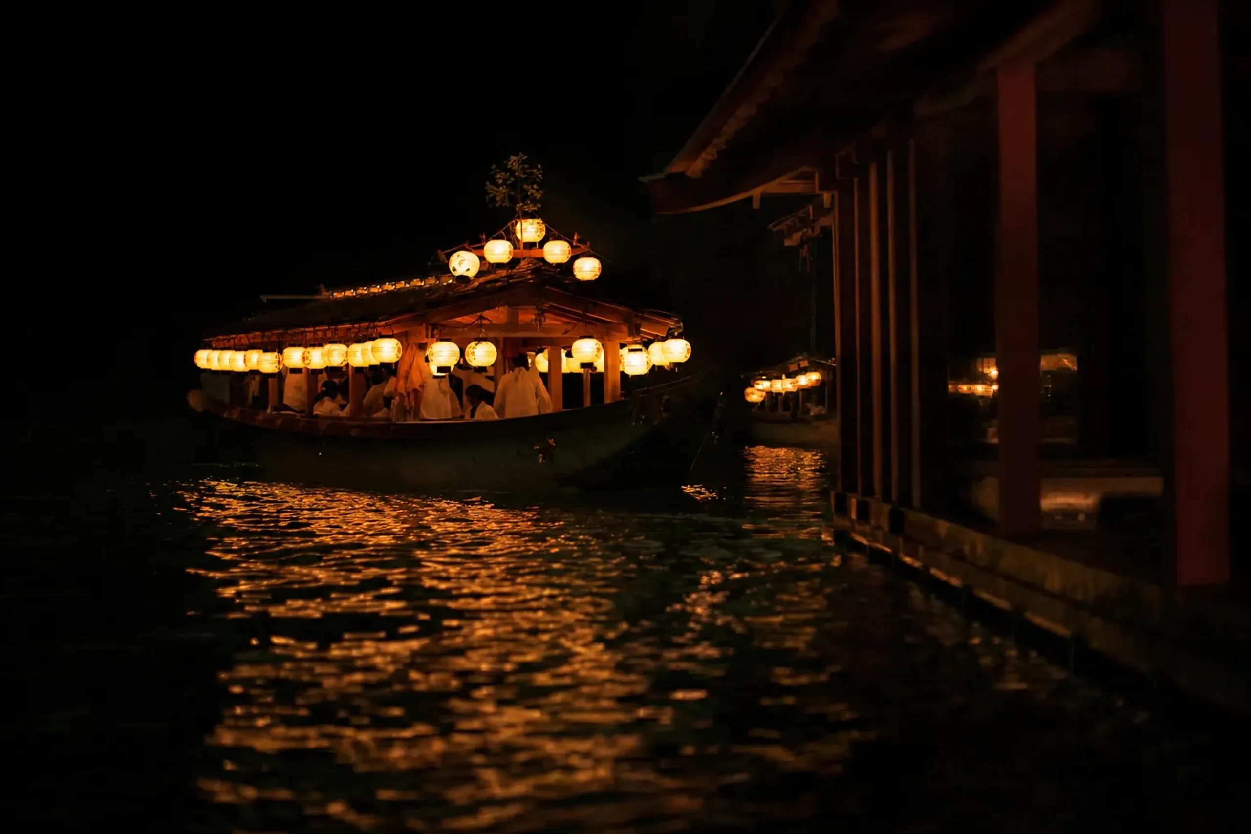 the returning Kangensai festival boat glowing with swinging paper lanterns reflected in the night sea near Itsukushima Shrine's corridor