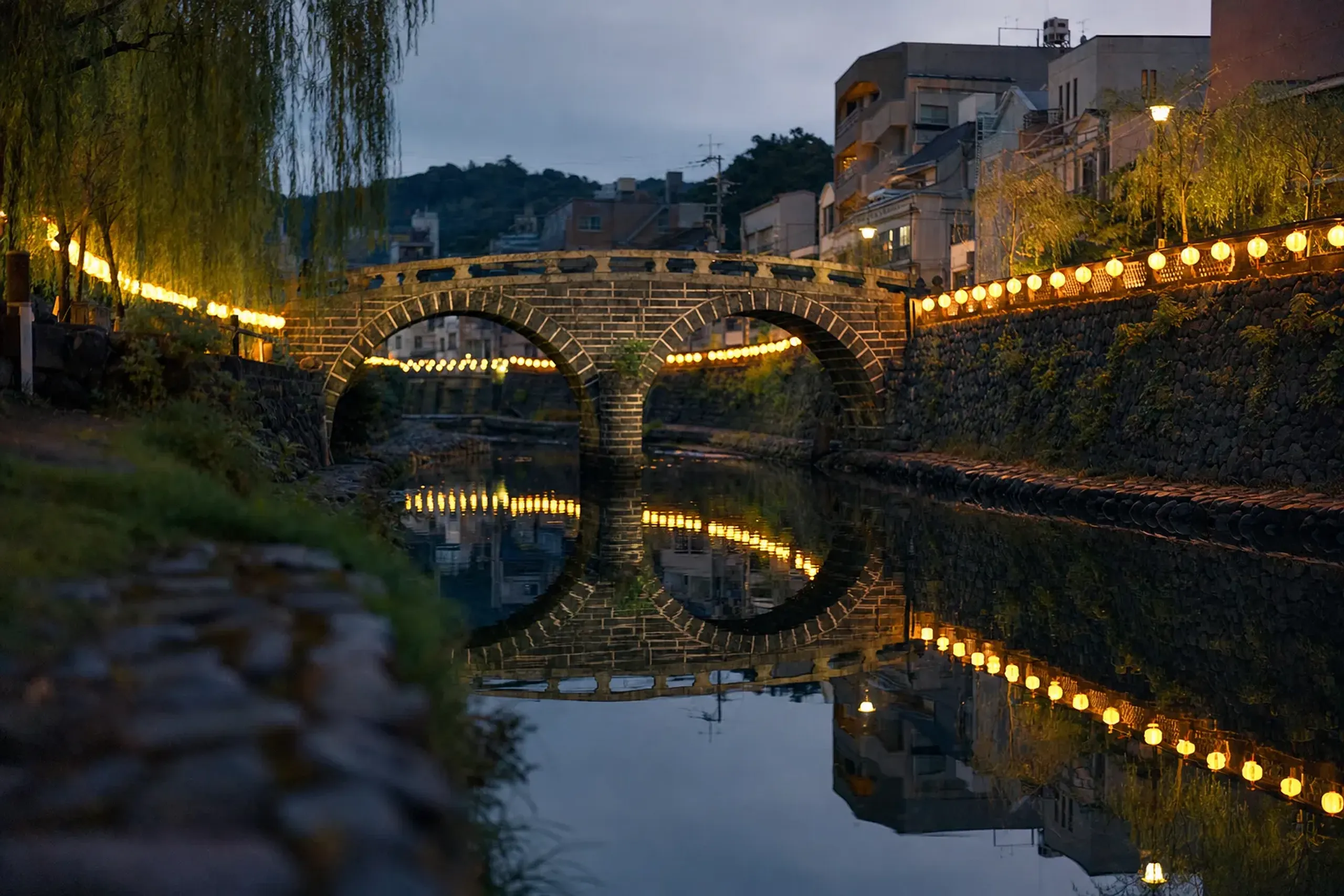 Yellow lanterns lining the banks of the Nakashima River, their reflections rippling in the dark water beside the double-arched stone Spectacles Bridge (Meganebashi)