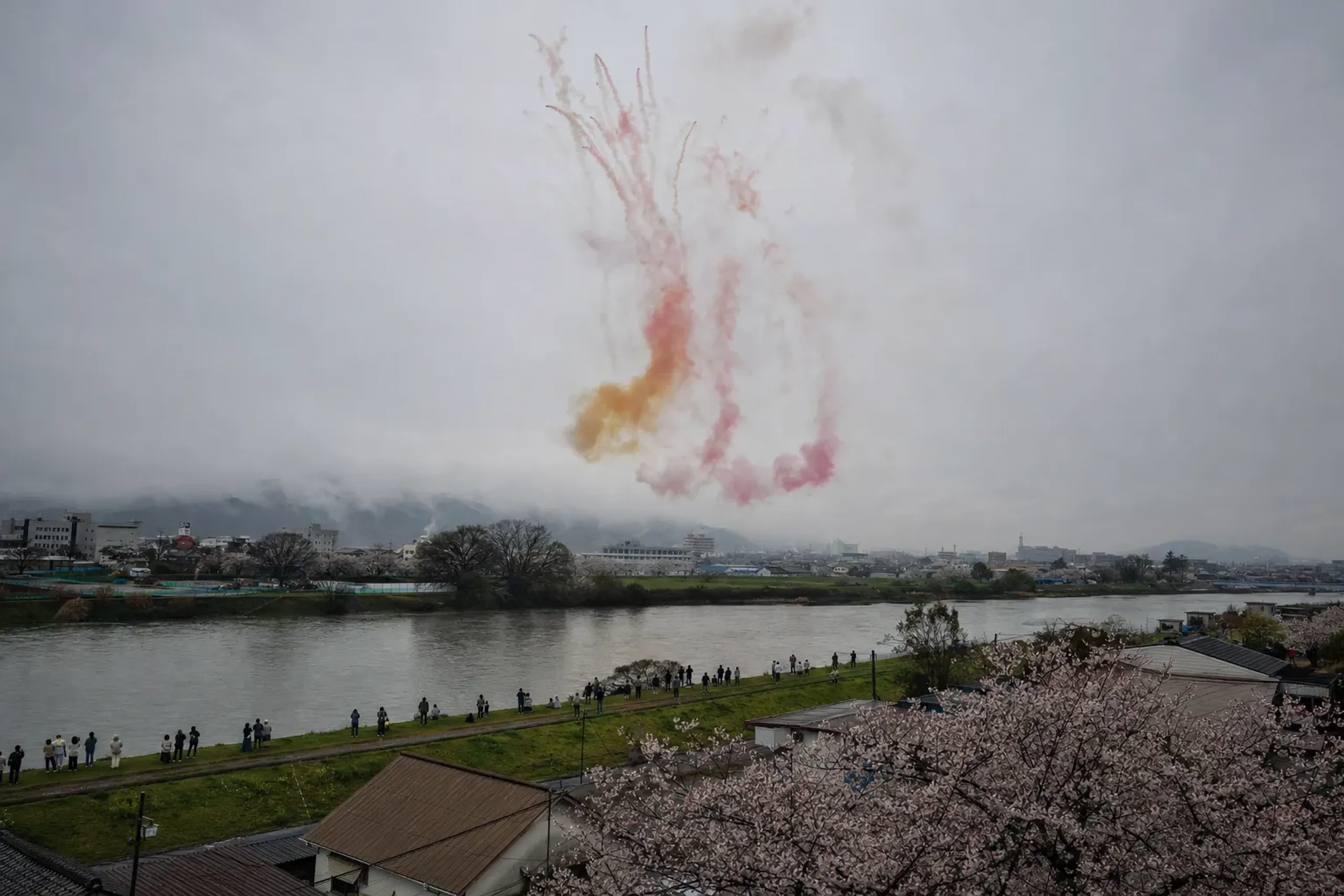 Coloured daytime smoke fireworks drifting over the Omono River against a pale sky at Omagari, Akita