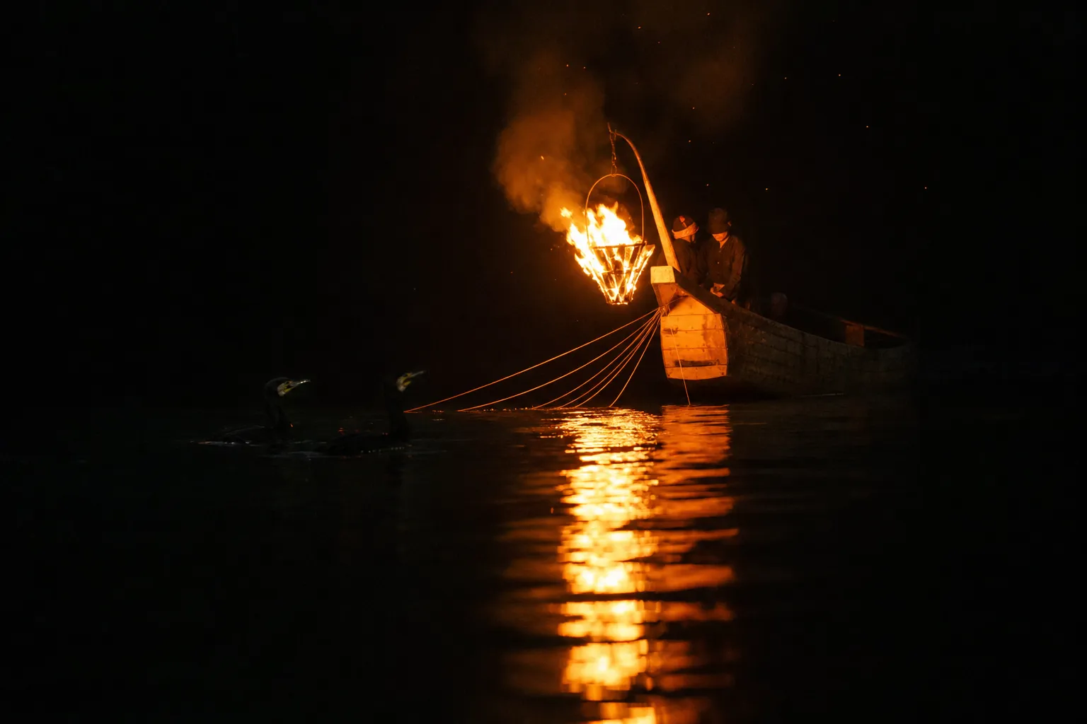 A low wooden ukai fishing boat on the dark Nagara River in Gifu, its bow fire basket casting amber torchlight across the mirror-still water, with lean black cormorant silhouettes surfacing near the re