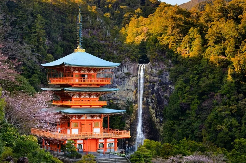 Five-story orange pagoda with a blue roof beside a cascading waterfall in a forested mountainside.