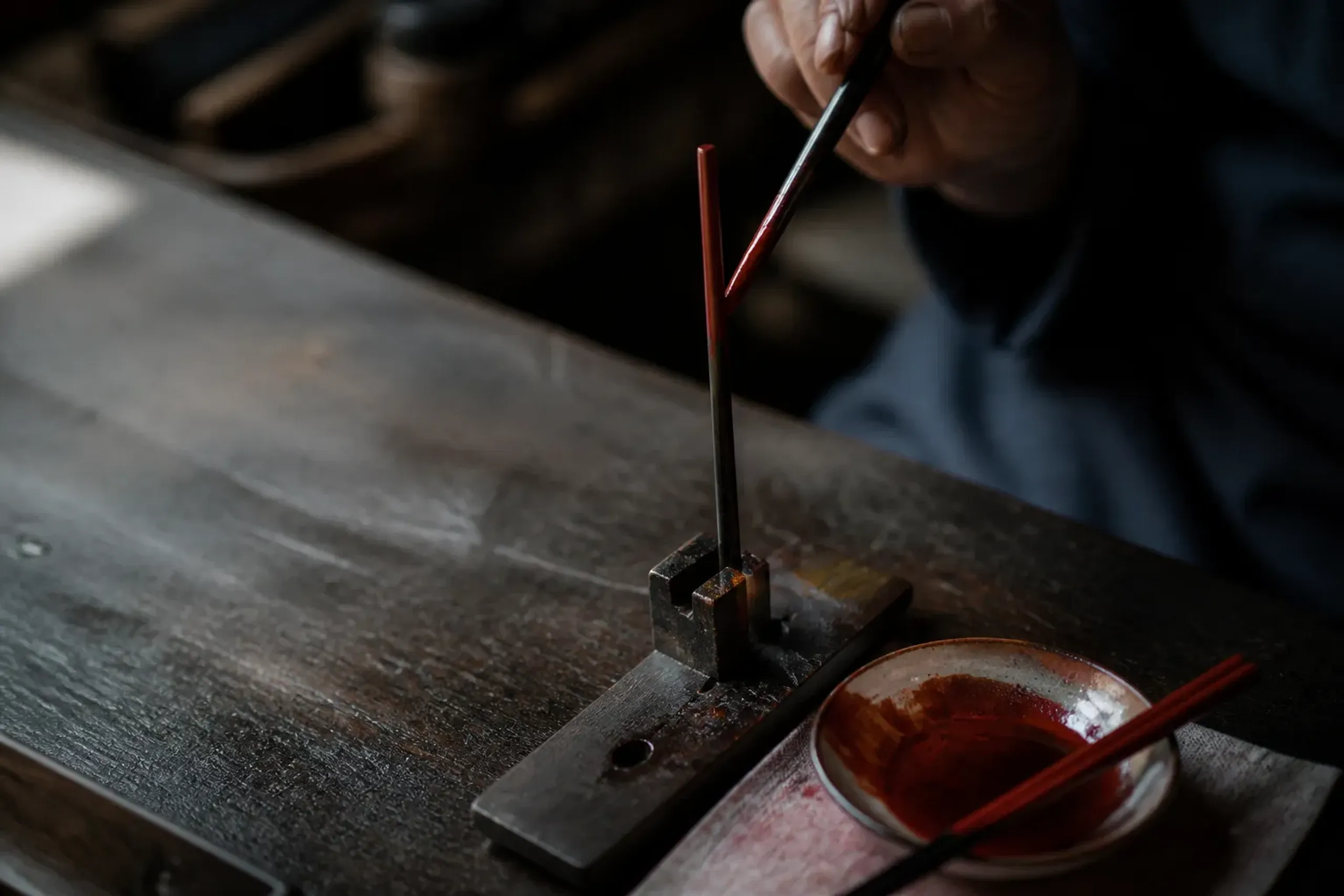 A pair of urushi chopsticks being decorated with coloured lacquer under an artisan's brush at a workshop bench in Takamatsu