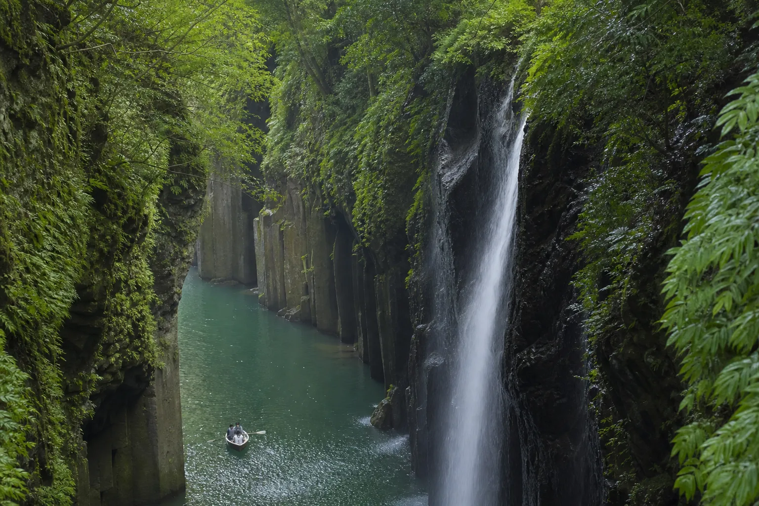 A lone rowboat drifting between the mossy basalt cliffs and waterfall of Takachiho Gorge