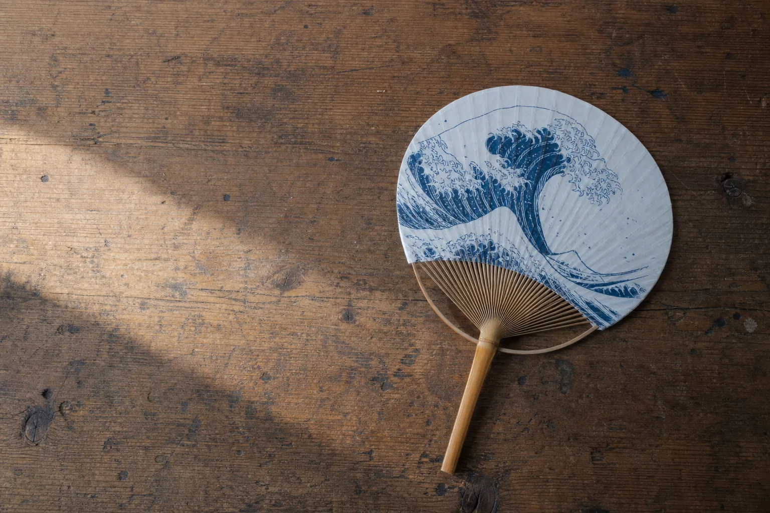 A finished bespoke Marugame uchiwa fan laid on a wooden table, its washi face hand-painted with a seasonal Seto Inland Sea wave motif, the single-piece split bamboo handle and forty radiating ribs vis