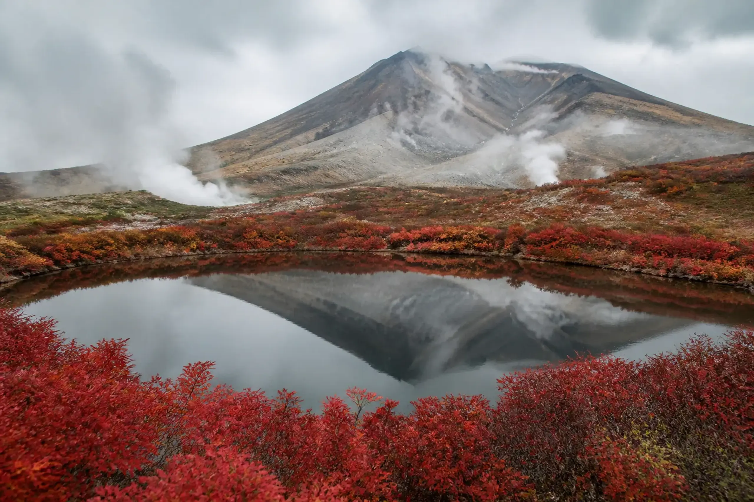 Steam rising from Sugatami Pond near Mount Asahidake surrounded by red autumn foliage.