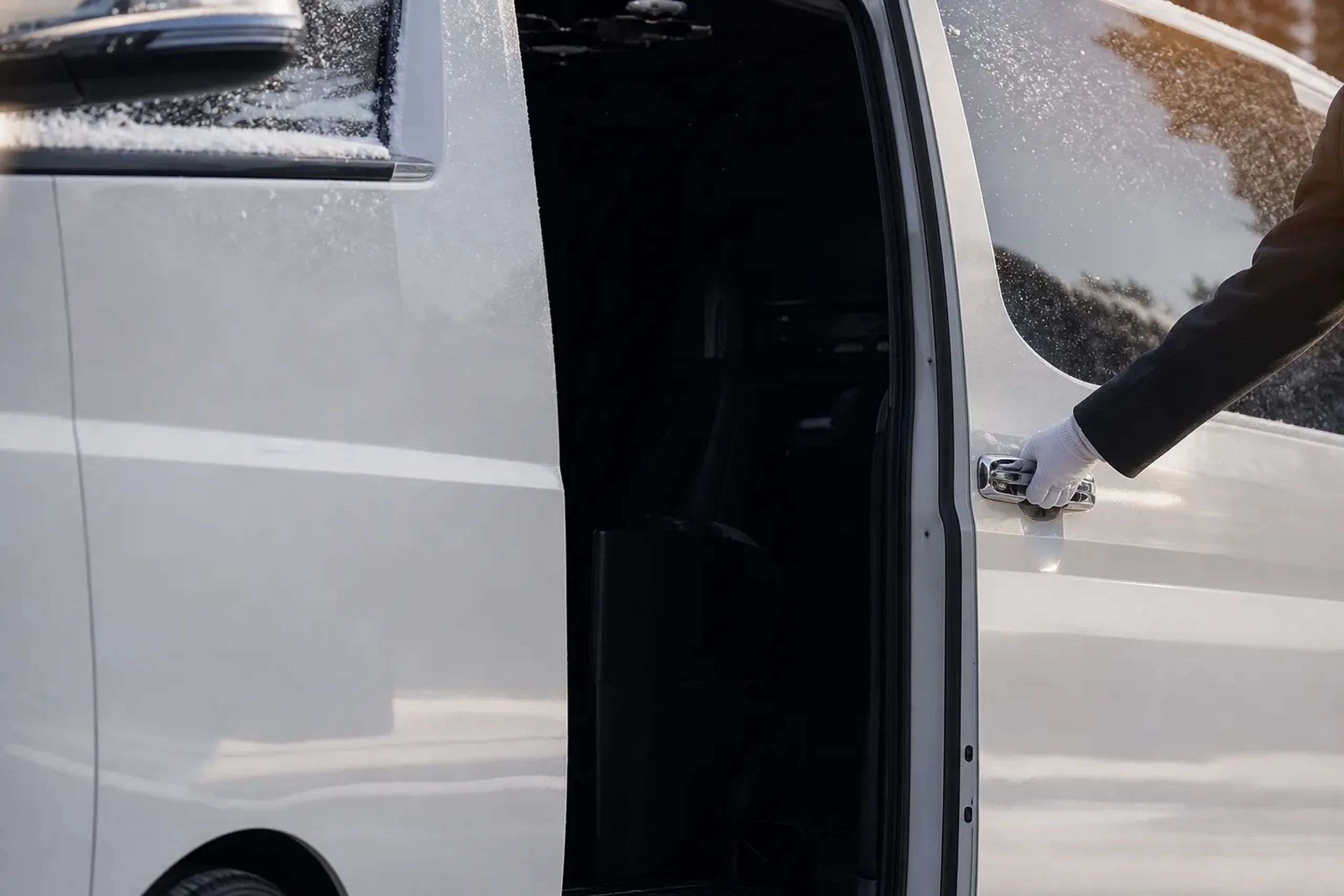 A dark-suited chauffeur's gloved hand holding open the sliding door of a white Toyota HiAce, no face visible