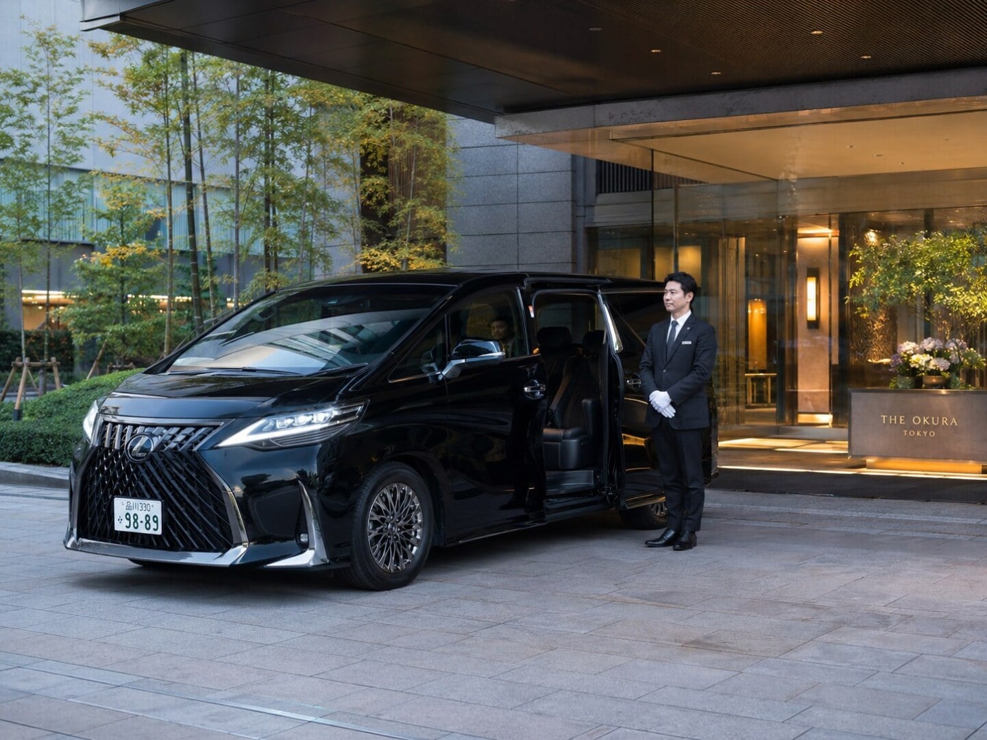 Chauffeur in a suit with white gloves standing by an open-door, black luxury van at The Okura Tokyo hotel entrance.