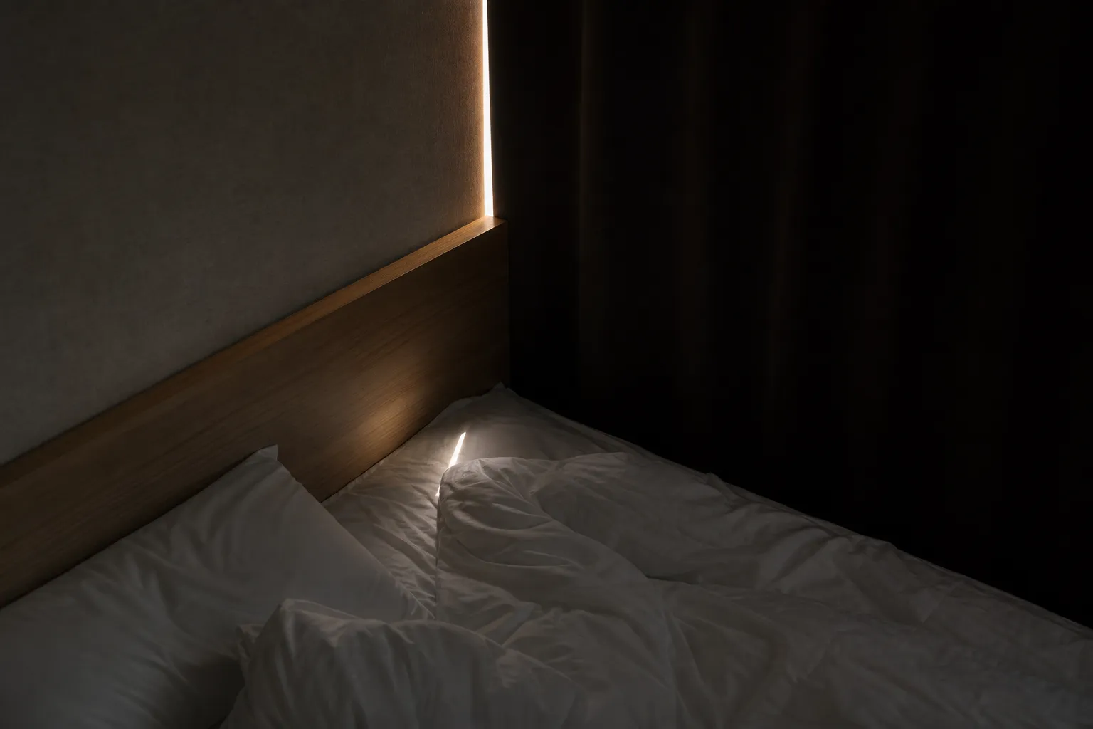 a blackout curtain edge leaking a thin blade of 5 a.m. light across a minimalist Tokyo hotel room's bed and wooden headboard
