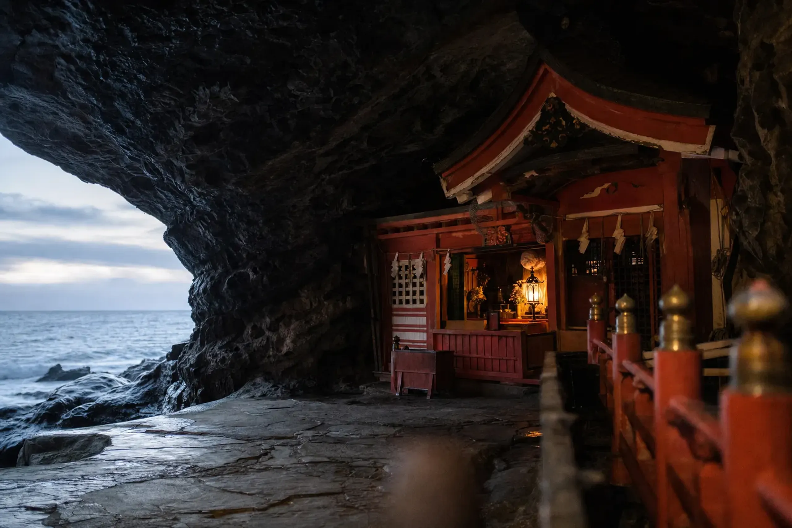 The vermillion-lacquered main hall of Udō Jingū set deep inside the dark wave-cut cave facing the Hyūga Sea, daylight glowing off the lacquer against black rock