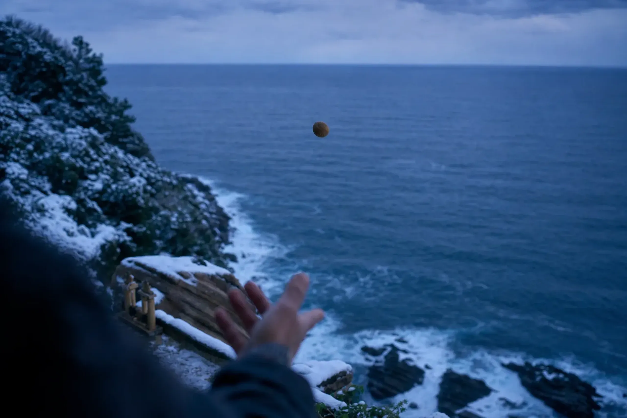 A small clay unda-nage ball arcing out over the cliff edge above the Hyūga Sea surf, caught mid-flight in the light