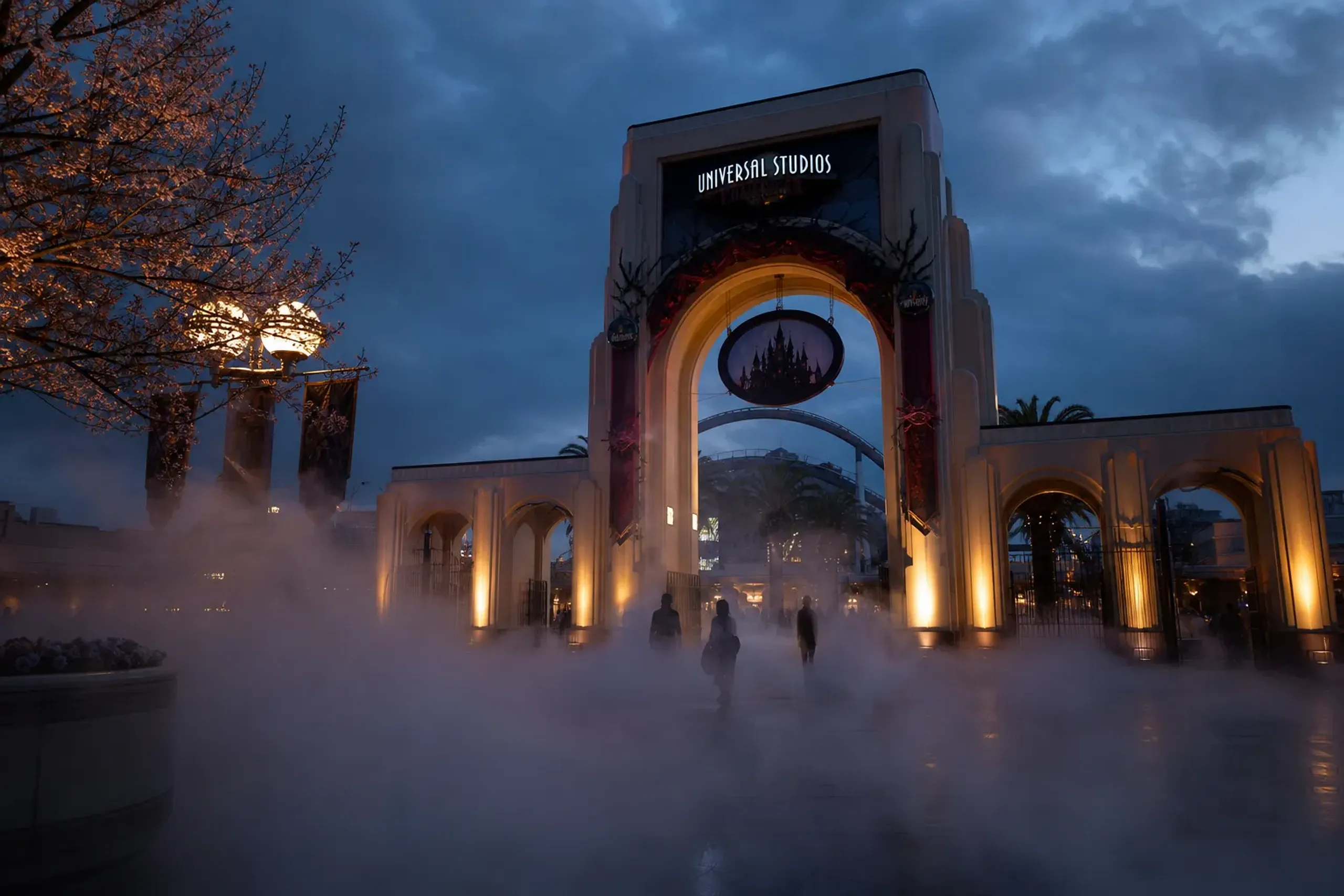 Fog machines exhaling low mist across the front gates of Universal Studios Japan at dusk, the sky the colour of cooled iron