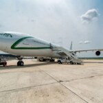 A wide-angle exterior view of a white Airbus A340-300 VIP aircraft parked on a concrete tarmac under a partly cloudy sky, featuring green accent stripes on the fuselage.