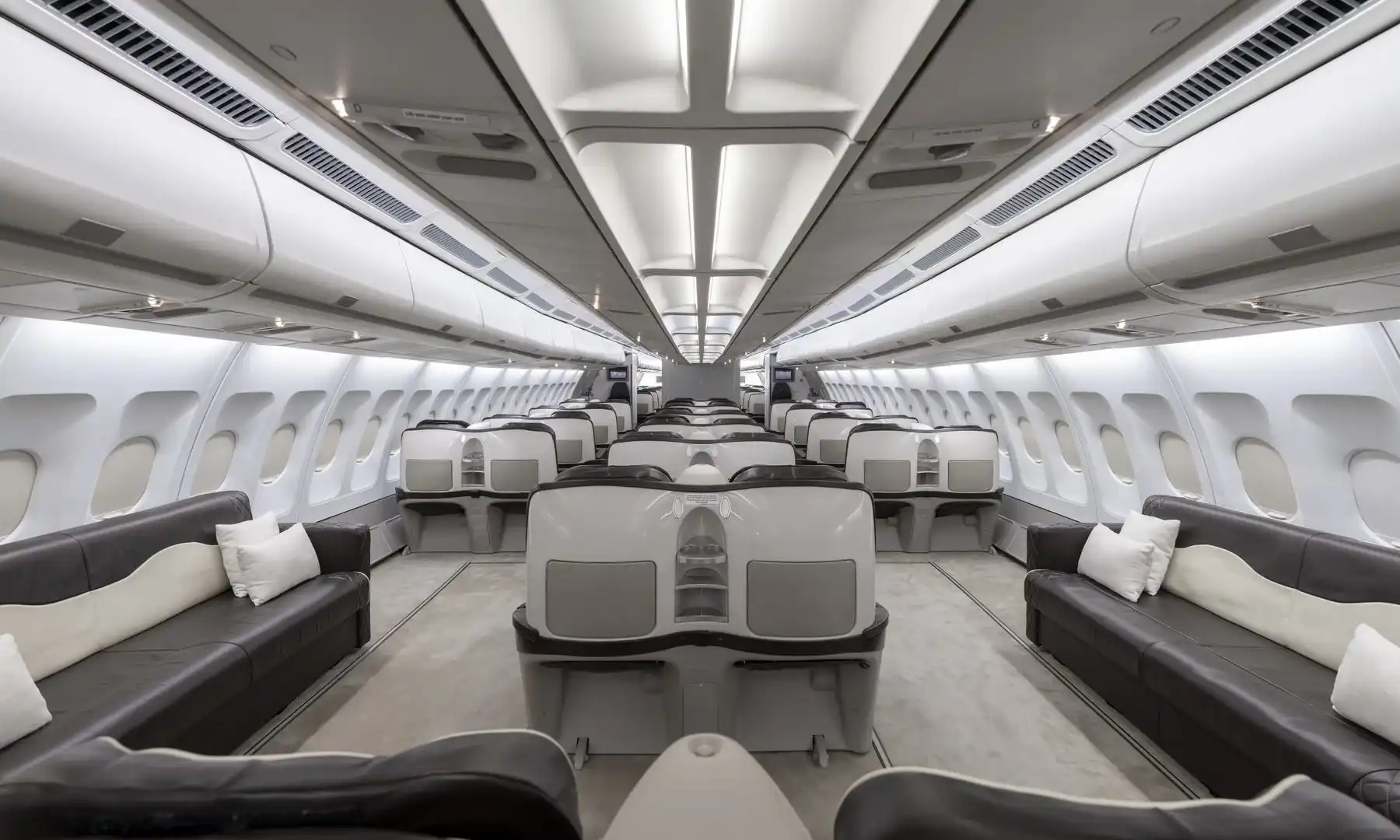 Interior view of a luxurious Airbus A340-300 VIP cabin, featuring long dark brown leather sofas with white pillows along the outer walls and premium, pod-style seats in the center aisle, illuminated by bright overhead lighting.