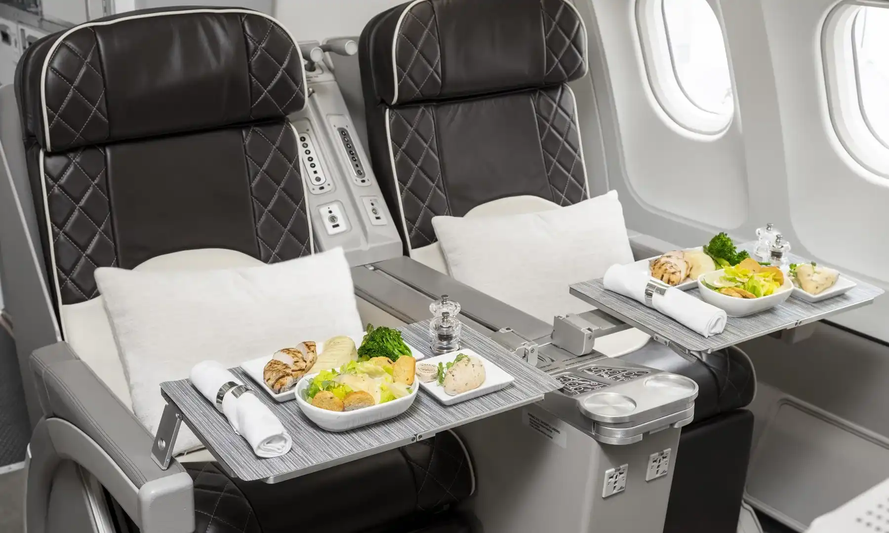 Close-up of two premium dark brown leather seats with diamond stitching next to a window in an Airbus A340-300. Deployed tray tables hold elegantly plated gourmet meals of chicken, salad, and vegetables, complete with silver napkin rings and salt and pepper shakers.