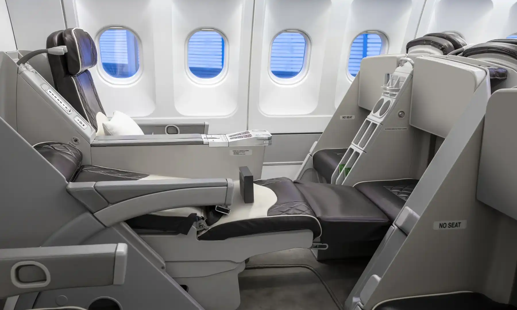 Side view of two dark brown leather seats with diamond stitching in an Airbus A340-300. The closest seat is fully reclined into a flat bed position, connecting with a padded footrest. Four cabin windows with blue shades drawn down are visible in the background.