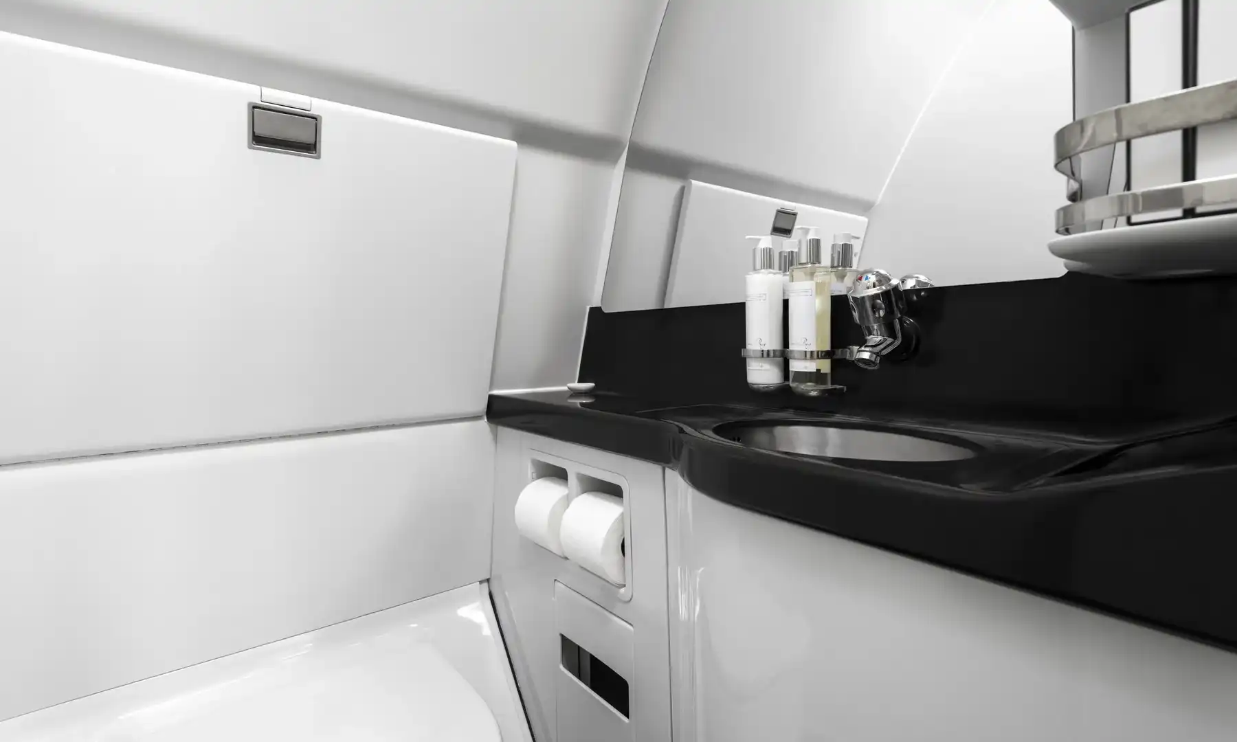 Interior view of an Airbus A340-300 VIP lavatory, featuring a modern design with clean white paneled walls, a glossy black countertop with an integrated stainless steel sink, a large mirror, and premium toiletries in a chrome dispenser.